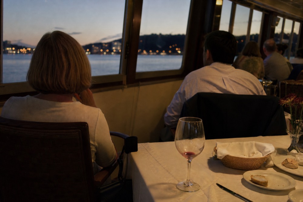 Diners seated at a table aboard a dinner cruise boat with harbor lights beyond the window, no text, no logos, no people