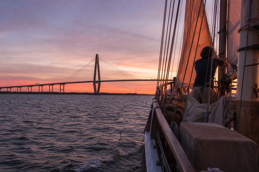 A tall ship sailing past the Ravenel Bridge at sunset in Charleston Harbor, no text, no logos, no people