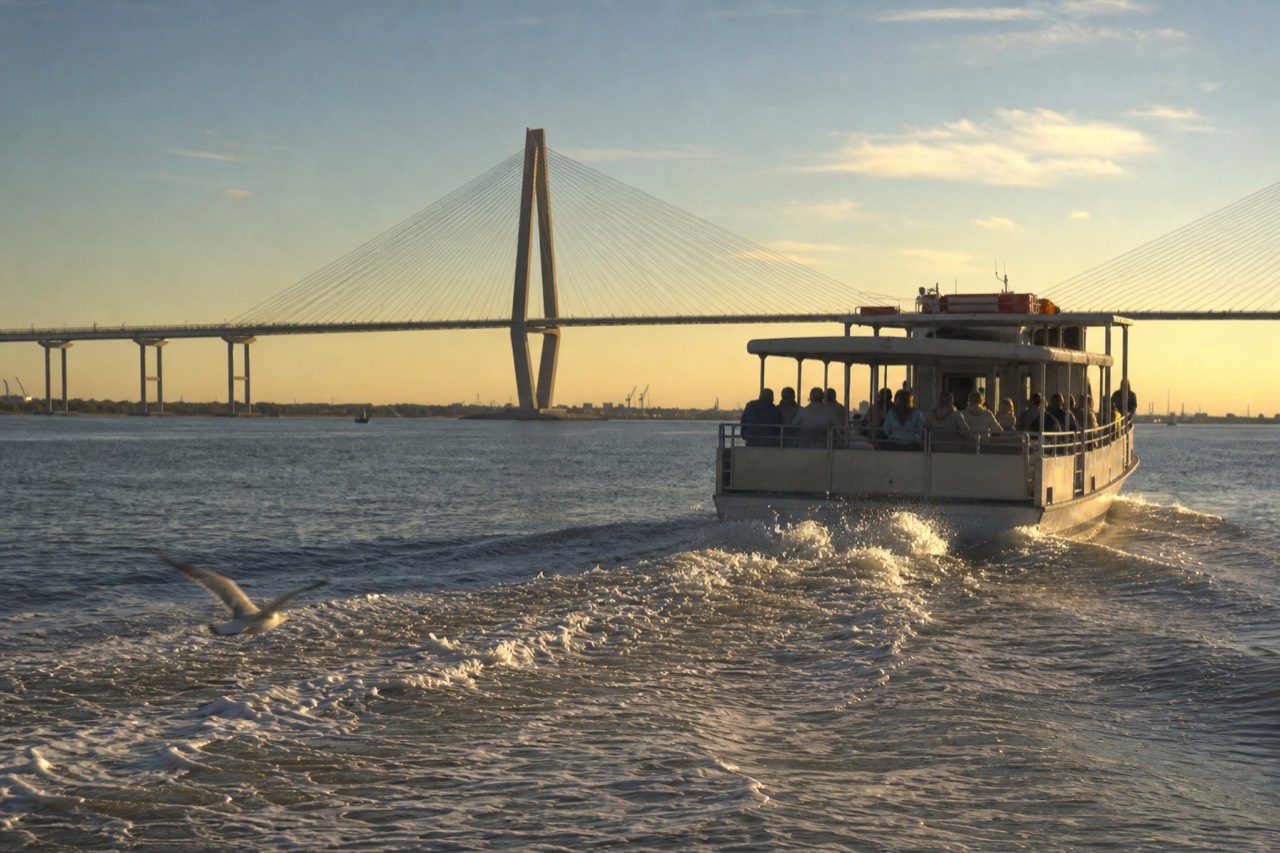 A tour boat crossing Charleston Harbor at golden hour with the Ravenel Bridge in the background, no text, no logos, no people