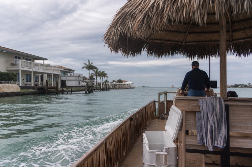 A tiki-style boat cruising past waterfront houses toward open harbor water, no text, no logos, no people