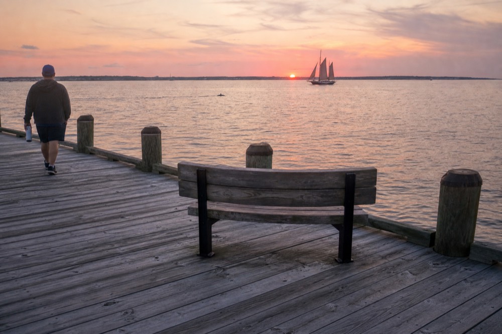 A schooner and a dolphin wake in the distance at sunset near a city shoreline, no text, no logos, no people