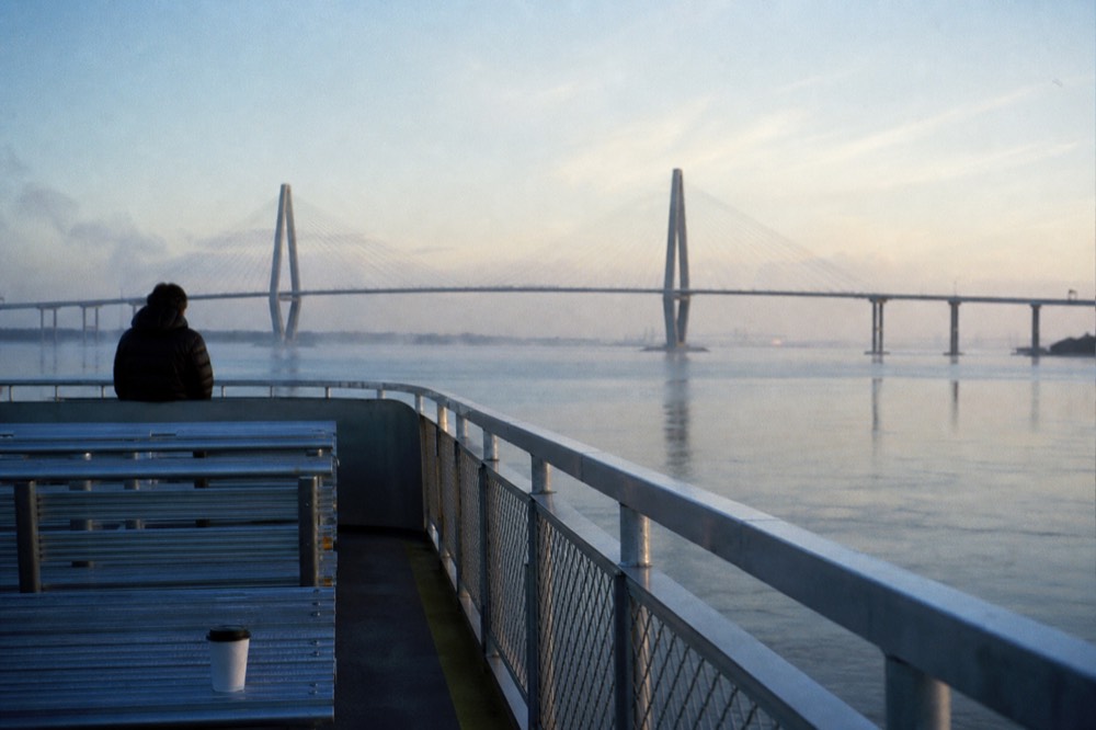 A quiet tour boat crossing calm harbor water toward the Ravenel Bridge in early morning light, no text, no logos, no people