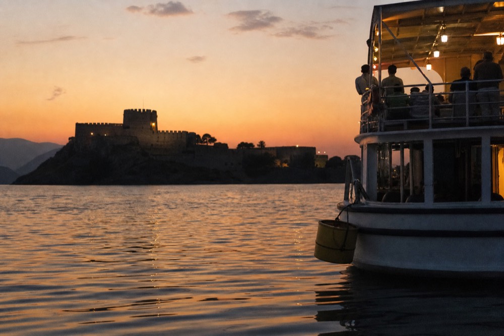 A cruise boat on calm harbor water with Fort Sumter silhouetted at sunset, no text, no logos, no people