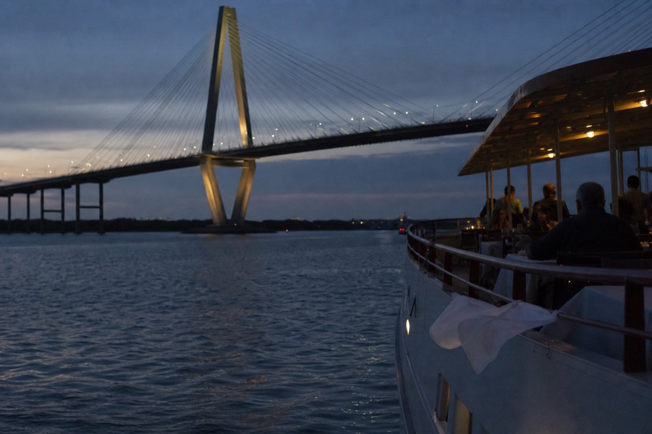 A dinner cruise boat on Charleston Harbor at dusk with the Ravenel Bridge in the background, no text, no logos, no people