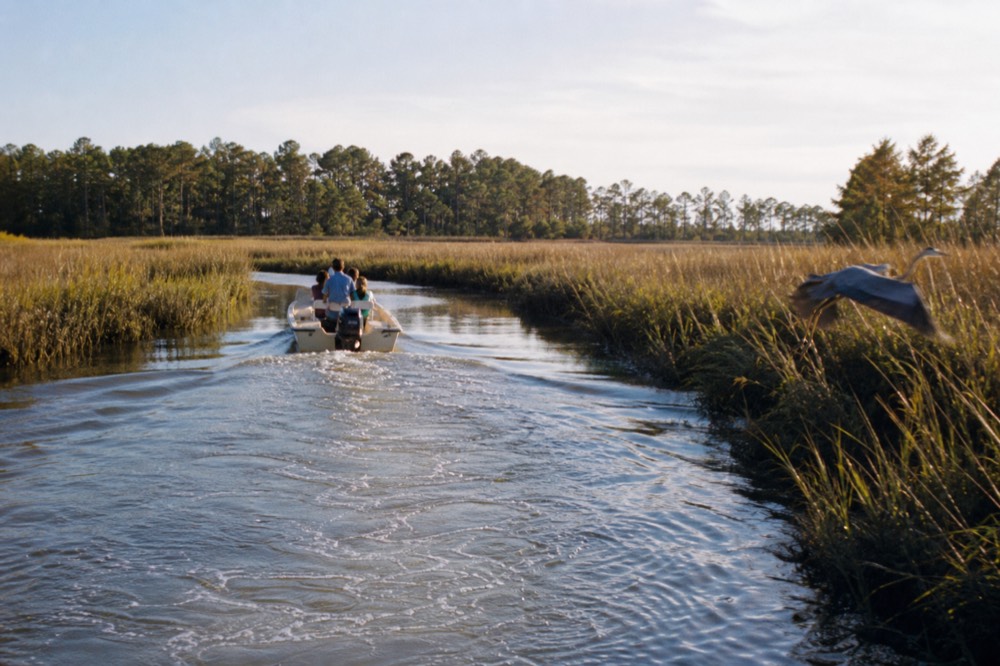 A small boat cruising a narrow tidal creek lined with marsh grass, no text, no logos, no people