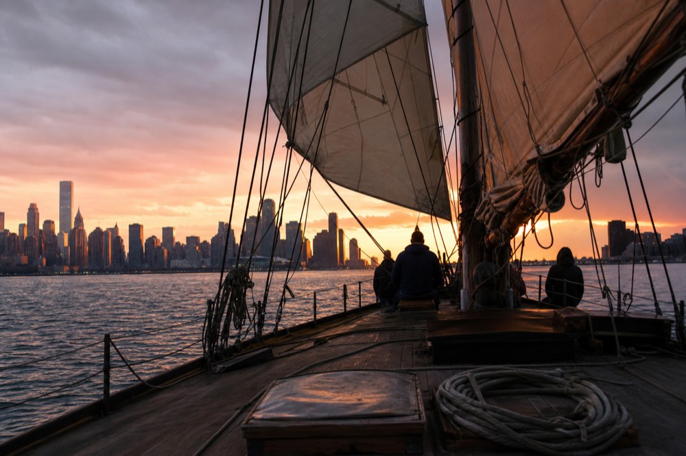 Schooner under sail on Charleston Harbor at sunset with the city skyline behind, no text, no logos, no people