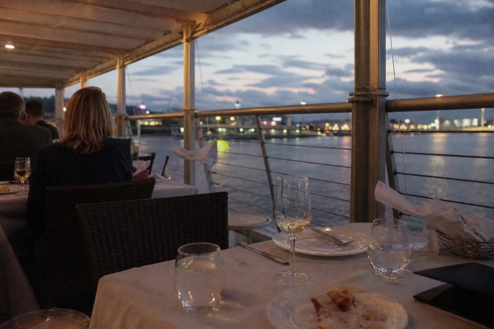 Set dinner table on a boat deck with harbor water and evening light through the windows, no text, no logos, no people