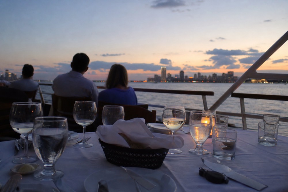 A set dinner table on a cruise ship deck with the city skyline beyond, no text, no logos, no people.