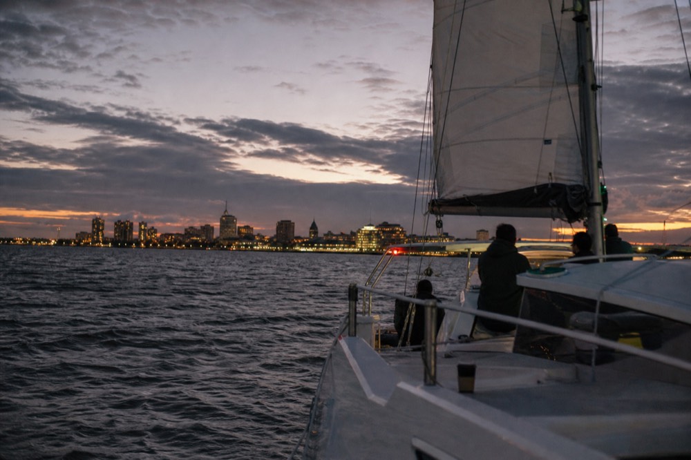 A catamaran under sail at dusk with the Charleston skyline glowing behind it, no text, no logos, no people