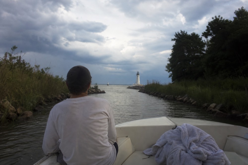 A small boat cruising a narrow creek toward open water near a lighthouse, no text, no logos, no people