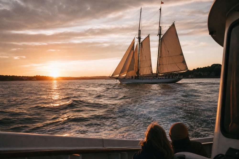 A tall schooner under full sail on Charleston Harbor at sunset, no text, no logos, no people.