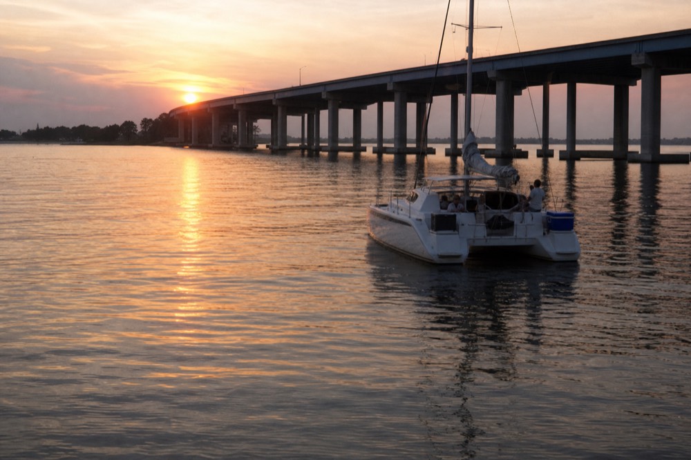 A catamaran sailboat on calm harbor water near the Ravenel Bridge at sunset, no text, no logos, no people