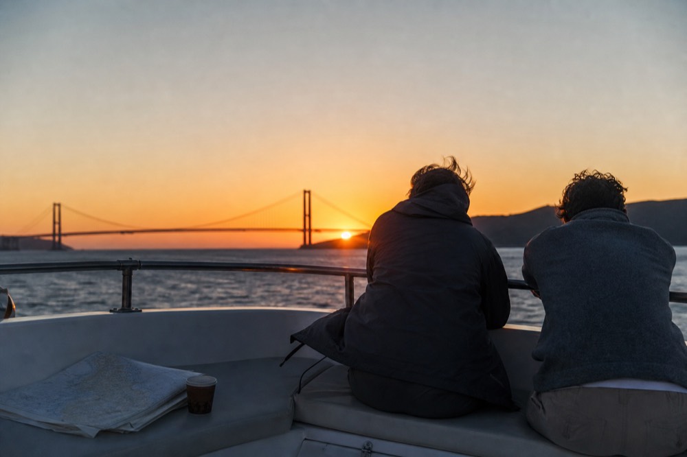 A couple at the bow rail of a boat watching the sun set behind the Ravenel Bridge, no text, no logos, no people